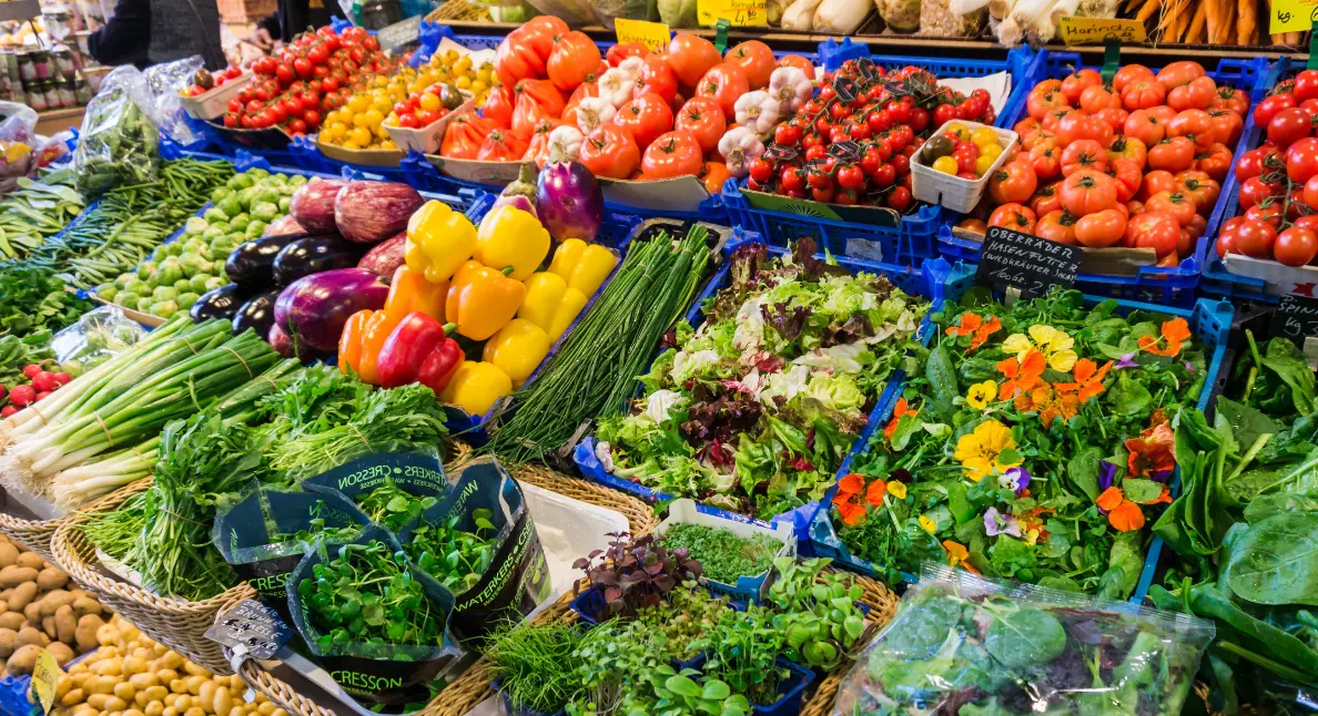 Buying local vegetables and greens at a vibrant market stall filled with peppers, tomatoes, lettuce, herbs, and edible flowers.