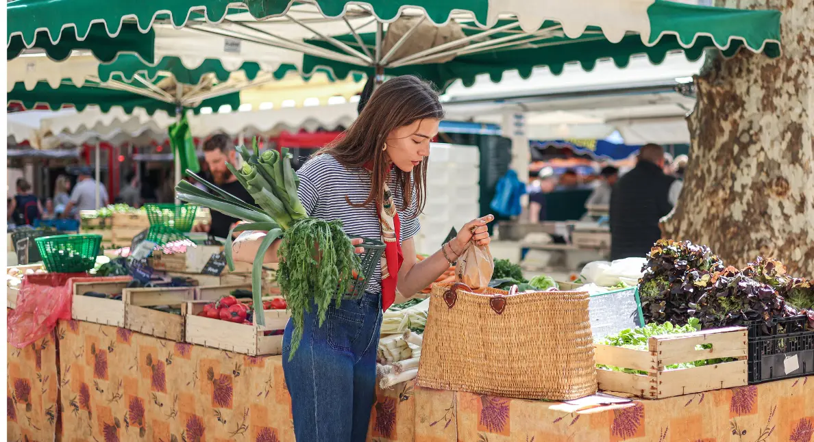 A woman packs fresh leeks and herbs into a wicker basket during a farmers market shopping trip under green canopy stalls.