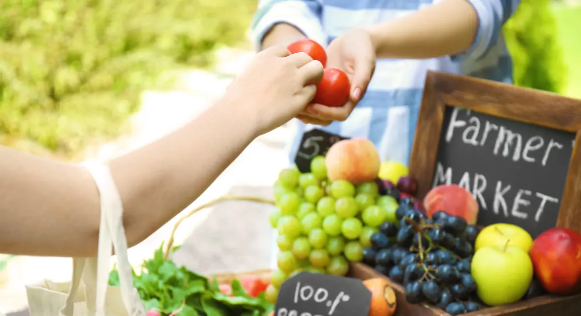 Local shopping at a farmers market with fresh tomatoes, grapes, apples, and greens being exchanged by hand.