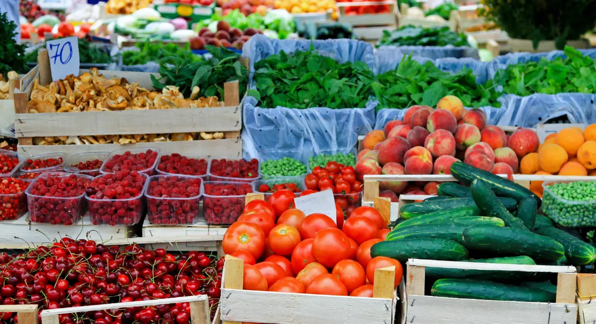 Produce market offering fresh tomatoes, cucumbers, berries, peaches, and leafy greens neatly arranged in wooden crates and baskets.