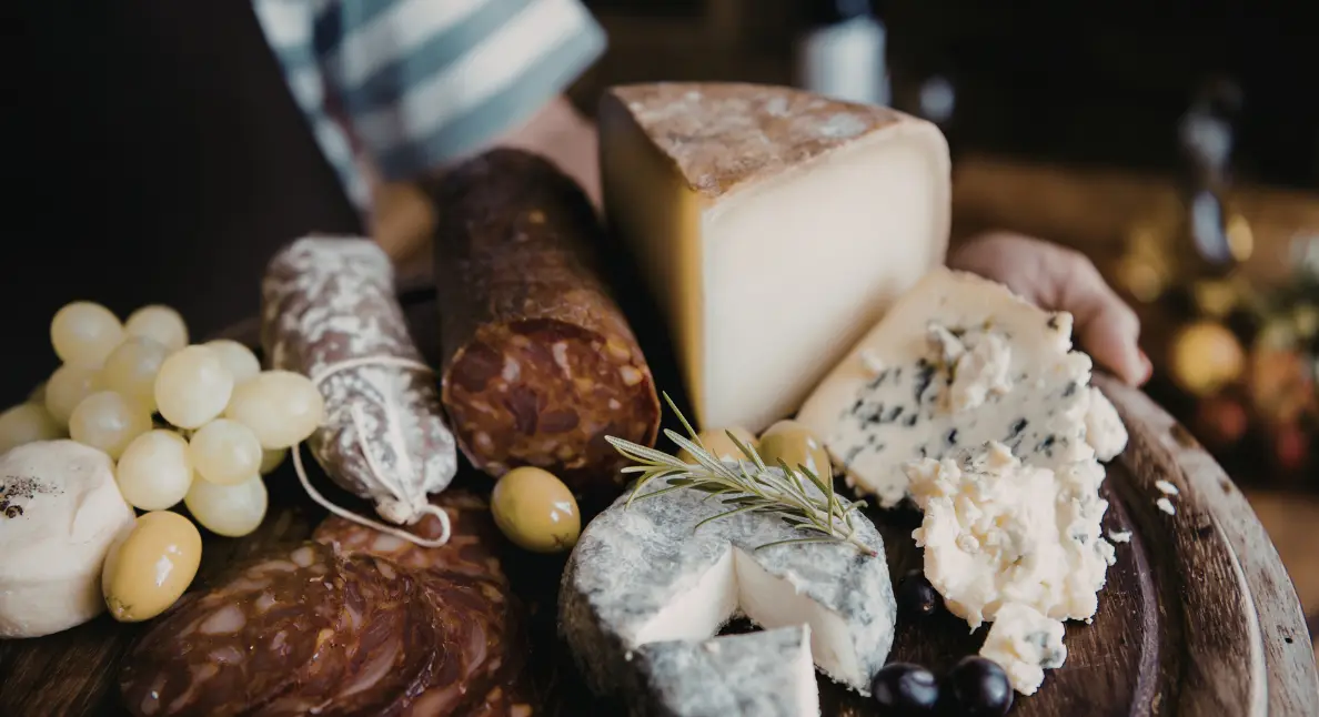 Small batch products including artisanal cheeses, cured meats, grapes, and olives arranged on a rustic wooden serving board.