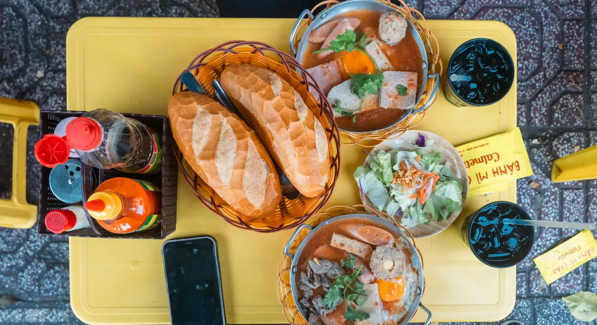 Authentic local food served on a yellow table featuring soup bowls, baguettes, salad, and condiments.