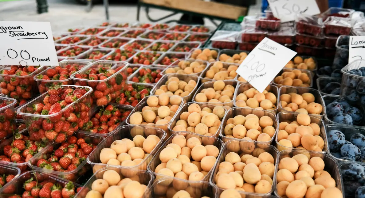 Farmers market display featuring strawberries, apricots, plums, and other fresh fruits neatly packed in containers with handwritten price signs.
