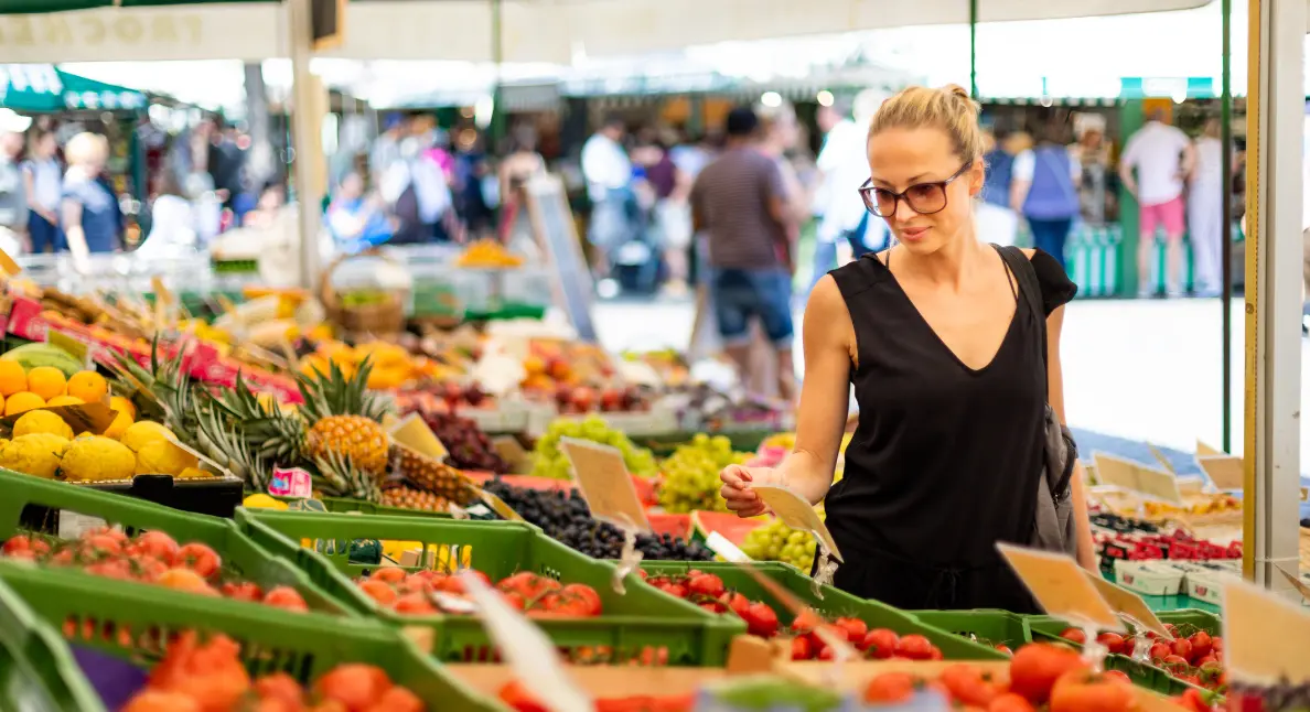 Fresh produce market scene with a woman browsing fruit and vegetables at an open air stall lined with green crates and price cards.