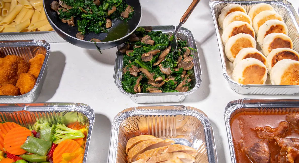 Handmade food products including sautéed greens, bread, and vegetables displayed in foil trays for ready meals.
