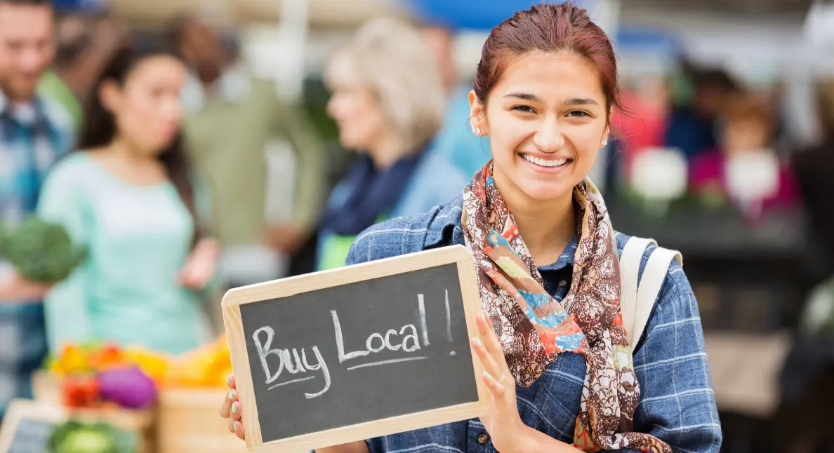 Local food creators promote community engagement with a smiling woman holding a "Buy Local!" sign at a farmers market.