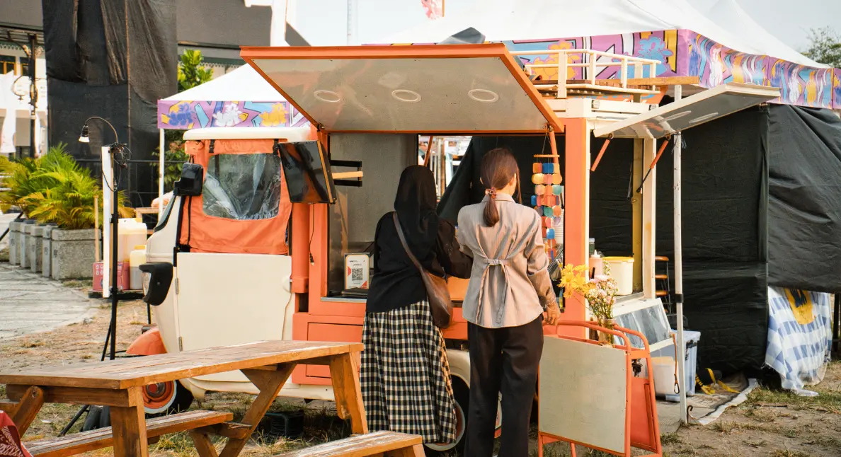 Local food dishes ordered from a vibrant mobile food cart at an outdoor event with two women waiting.