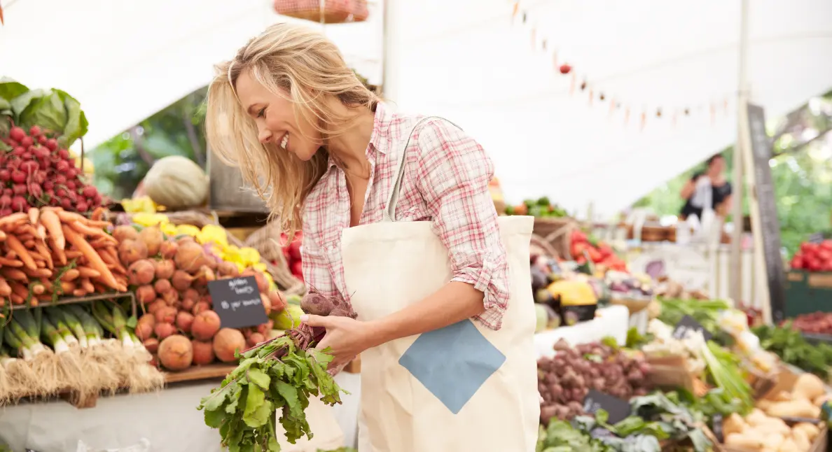 Local food movement represented by a smiling woman choosing beets at a farmers market stall filled with root vegetables.