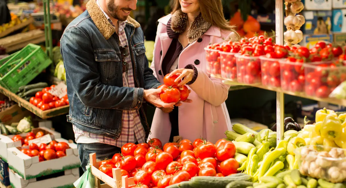 Produce market shopper selecting a ripe tomato while holding a wicker basket of fresh fruit beside wooden produce bins.