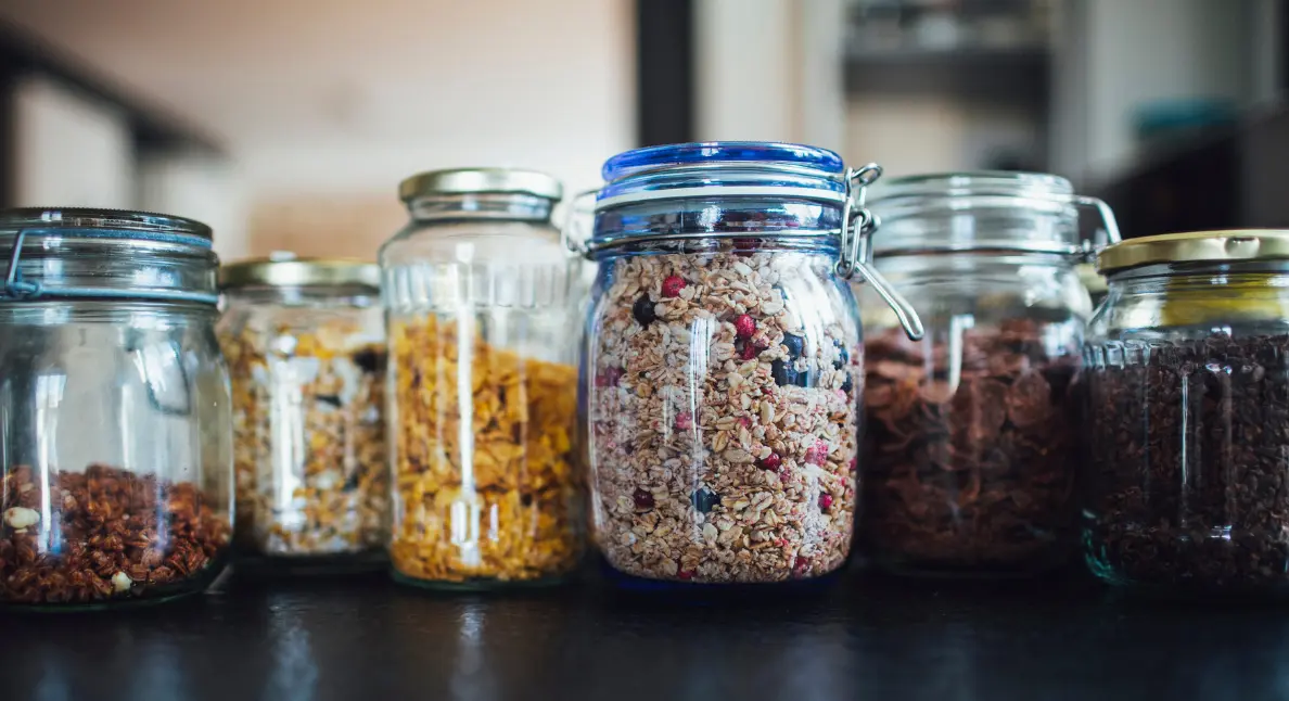 Premium food products stored in clear glass jars, including granola, cereal flakes, and dried ingredients arranged neatly on a kitchen counter.