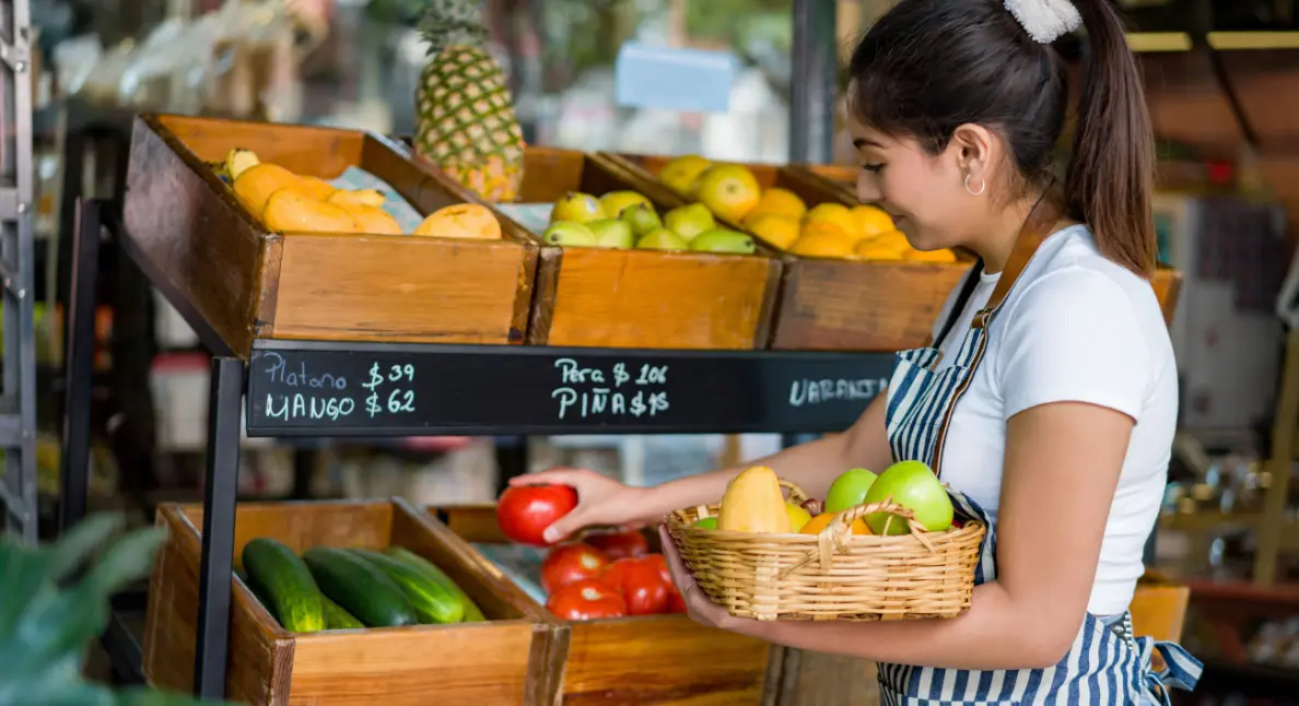 Produce market shopper selecting a ripe tomato while holding a wicker basket of fresh fruit beside wooden produce bins.
