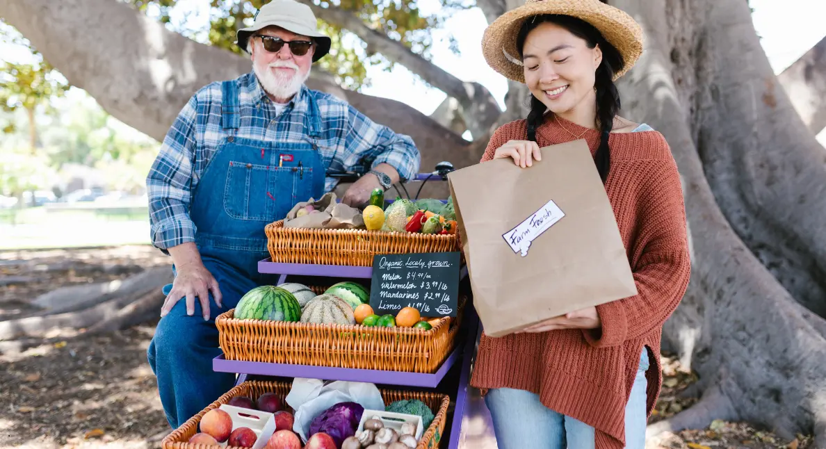 A smiling woman holds a farm fresh bag while buying from farmers at a charming outdoor produce cart.