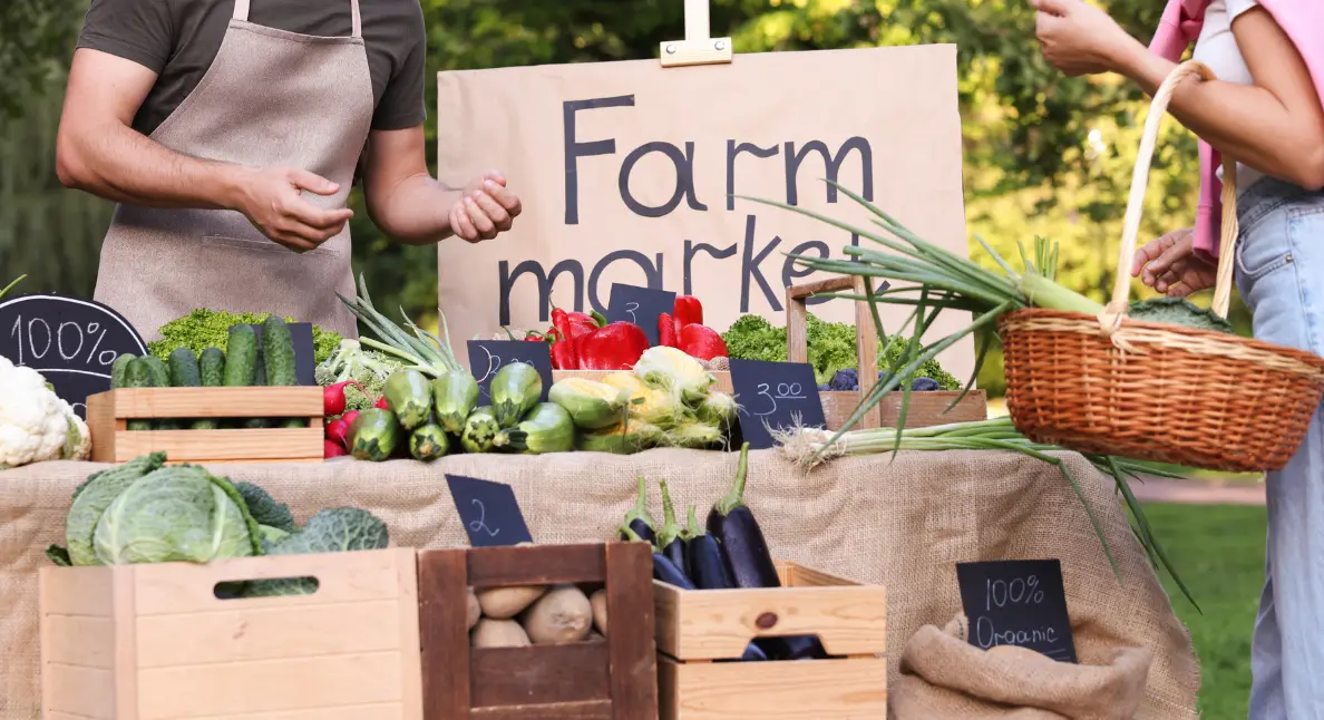 A vendor and customer browse a farm market stall stocked with 100% organic vegetables, embodying the spirit of buying local.