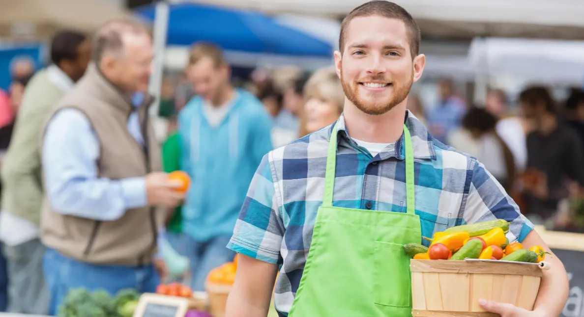 A smiling vendor in a green apron holds a basket of mixed vegetables, encouraging buying local at a busy farmers market. A smiling vendor in a green apron holds a basket of mixed vegetables, encouraging buying local at a busy farmers market.