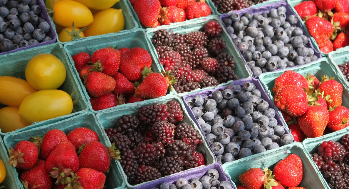 Pints of organic fruits including strawberries, blueberries, and blackberries displayed in green cartons at a market stall.