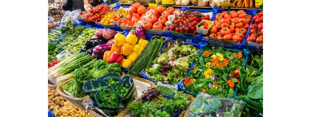 Buying local vegetables and greens at a vibrant market stall filled with peppers, tomatoes, lettuce, herbs, and edible flowers.