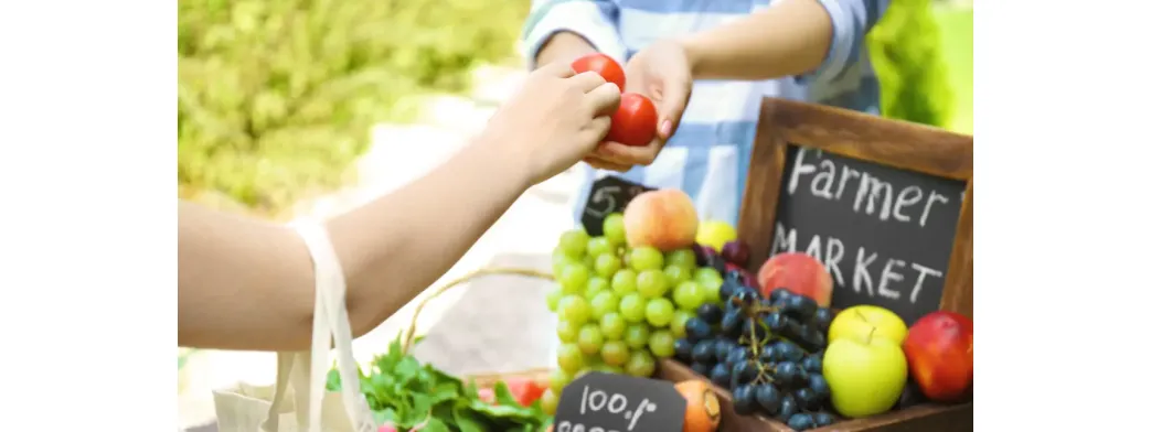 Local shopping at a farmers market with fresh tomatoes, grapes, apples, and greens being exchanged by hand.