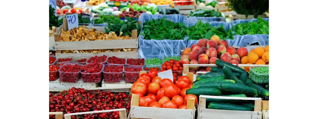 Produce market offering fresh tomatoes, cucumbers, berries, peaches, and leafy greens neatly arranged in wooden crates and baskets.