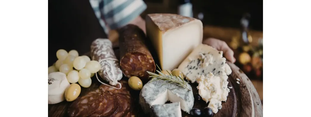 Small batch products including artisanal cheeses, cured meats, grapes, and olives arranged on a rustic wooden serving board.