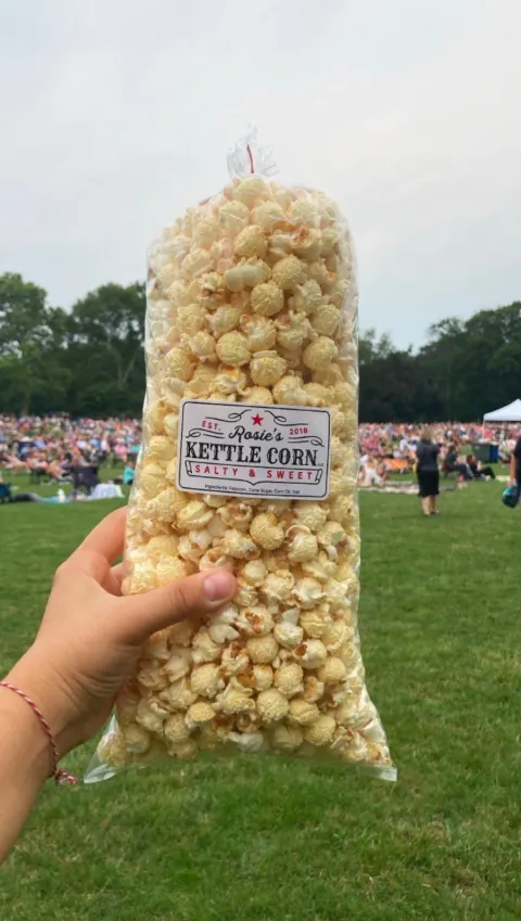 Hand holding a bag of Rosie’s Kettle Corn salty and sweet popcorn at an outdoor festival on a grassy field.