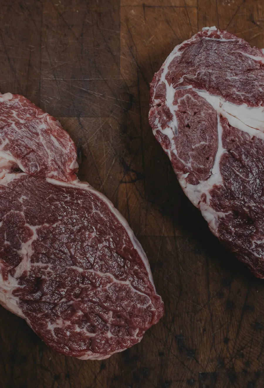 Meat cuts of marbled raw beef displayed on a wooden butcher block surface.