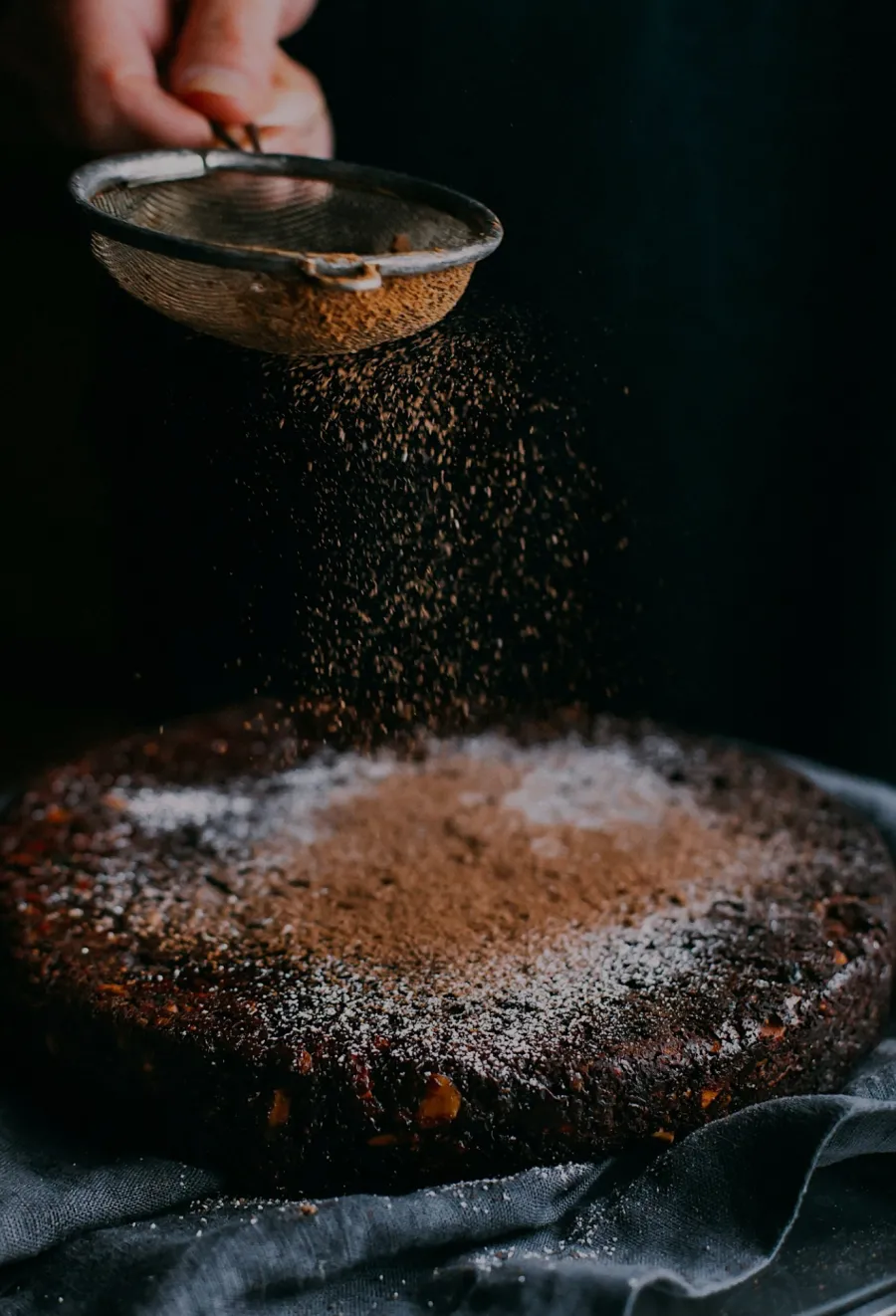 Chocolate cake being dusted with cocoa powder using a sieve over a dark background.