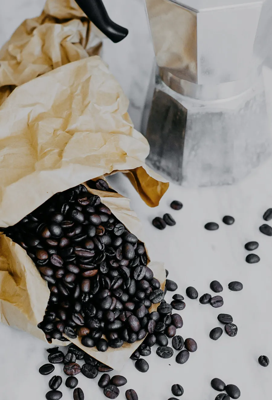 Coffee beans spilling from a paper bag beside a moka pot on a white marble countertop.