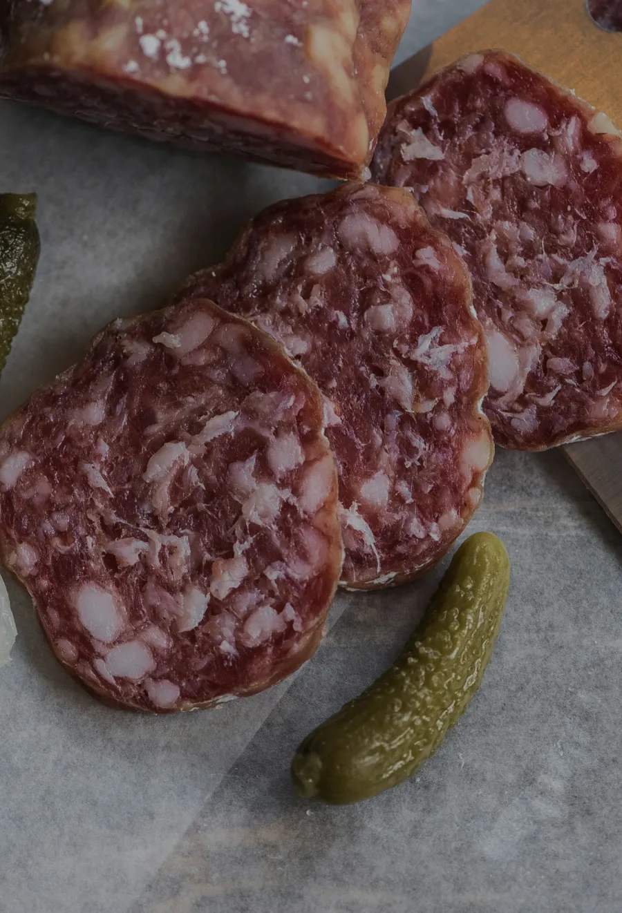Salami slices arranged on parchment paper with whole pickles and a partially cut salami log.