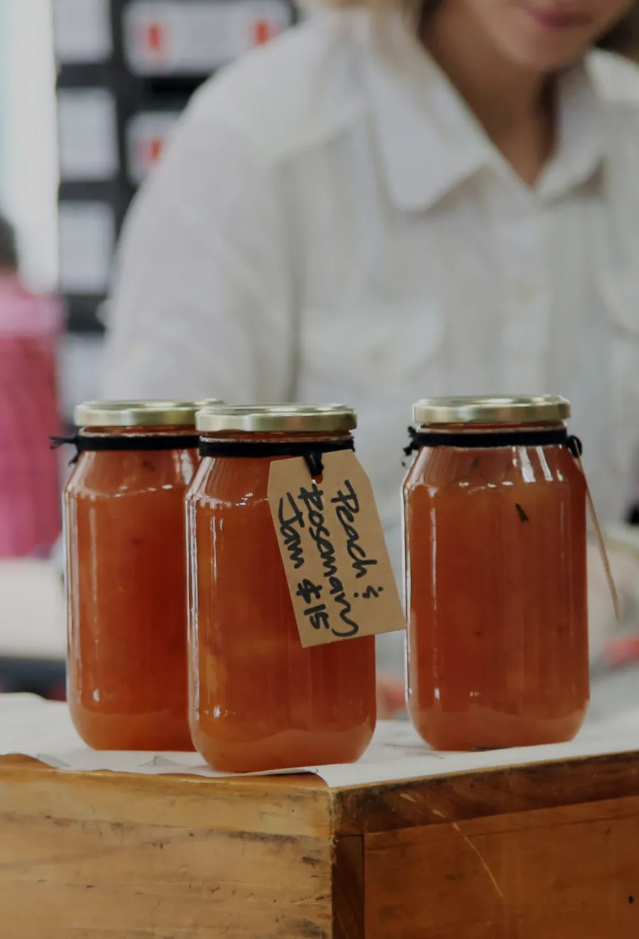 Peach jam jars with handwritten labels displayed at a market stall with a vendor in the background.