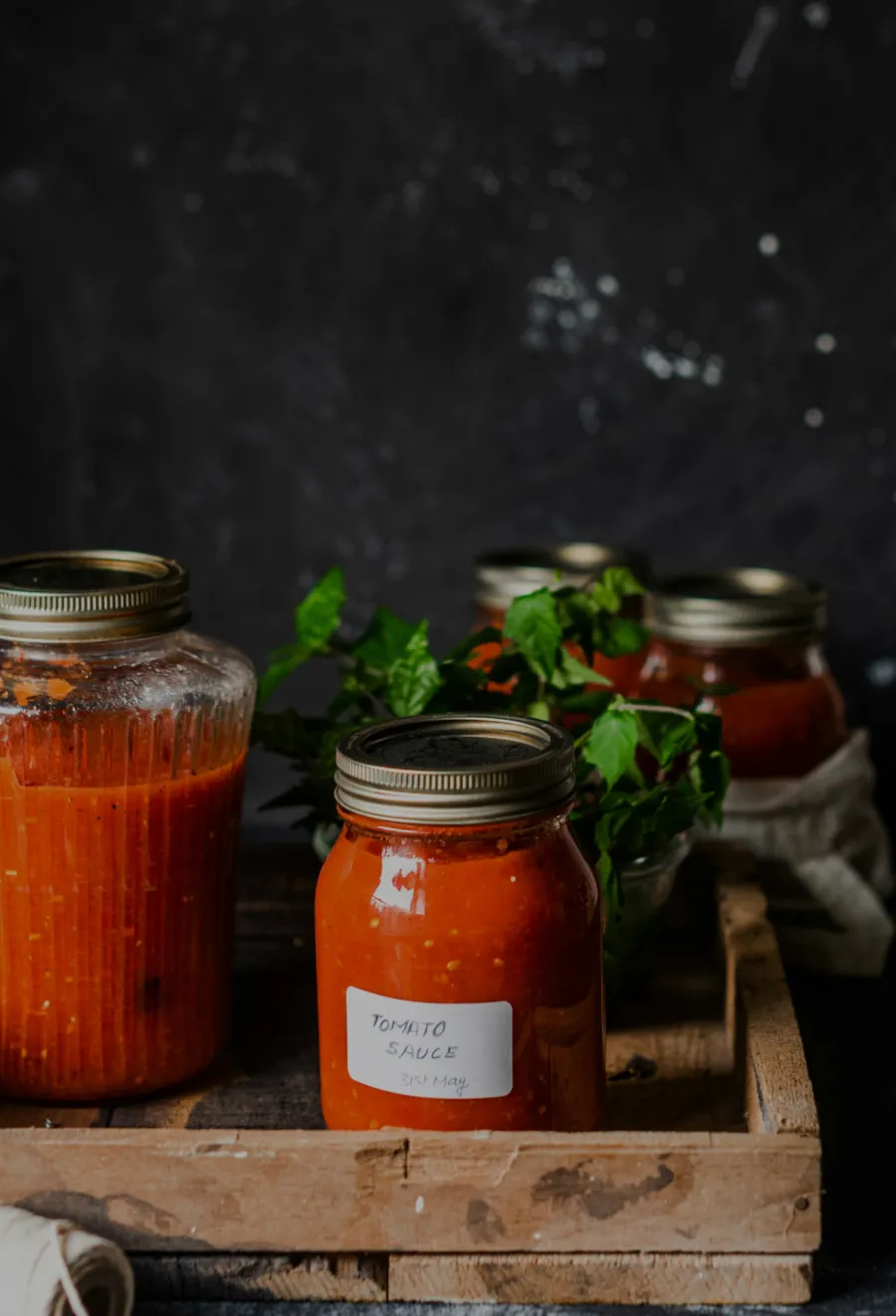 Tomato sauce in labeled mason jars placed in a rustic wooden crate with fresh basil in the background.