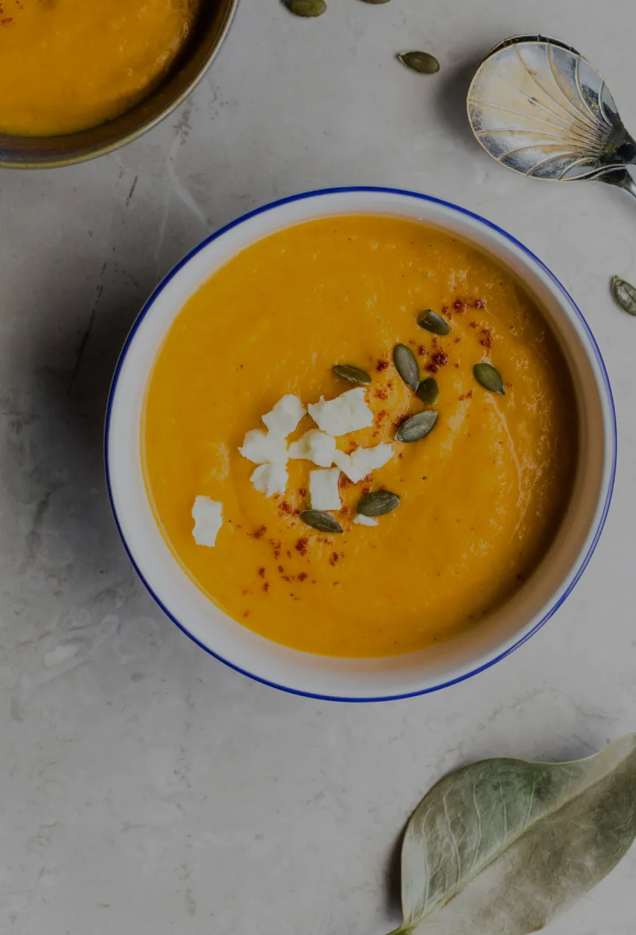 Cream soup served in a white bowl, topped with feta cheese, paprika, and pumpkin seeds on a light surface.