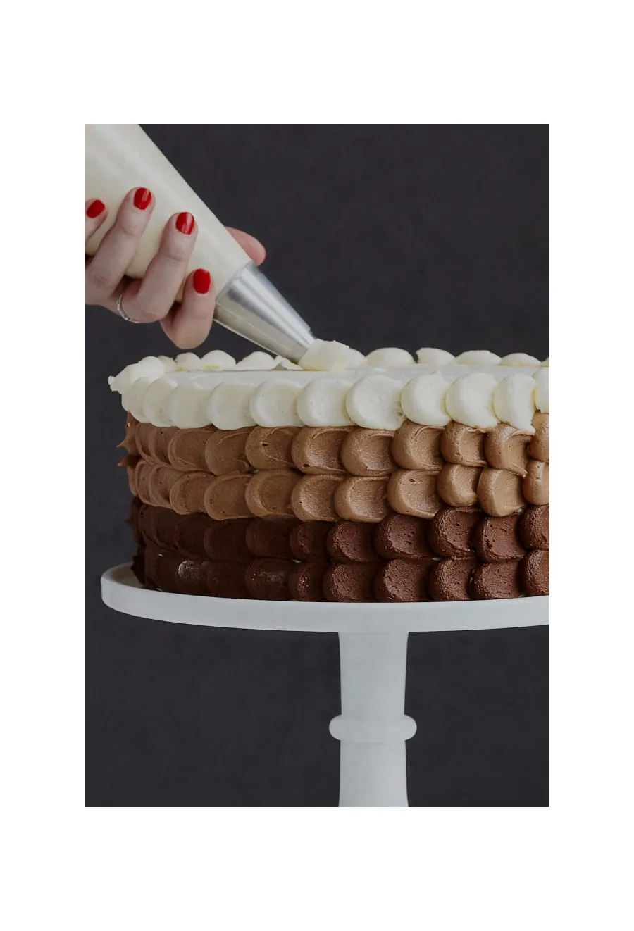Baked goods being decorated with vanilla frosting over layers of chocolate ombré icing on a cake stand.