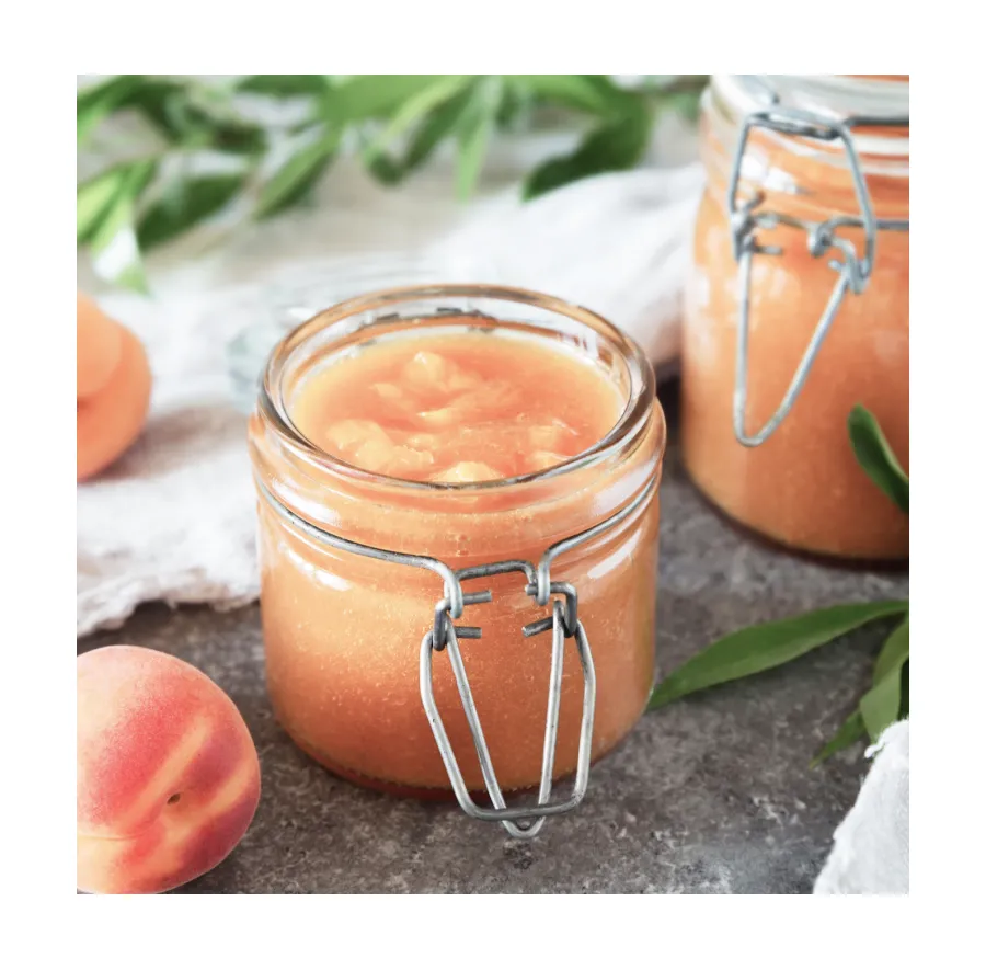 Peach preserves in a glass jar with fresh peaches and greenery on a stone countertop.